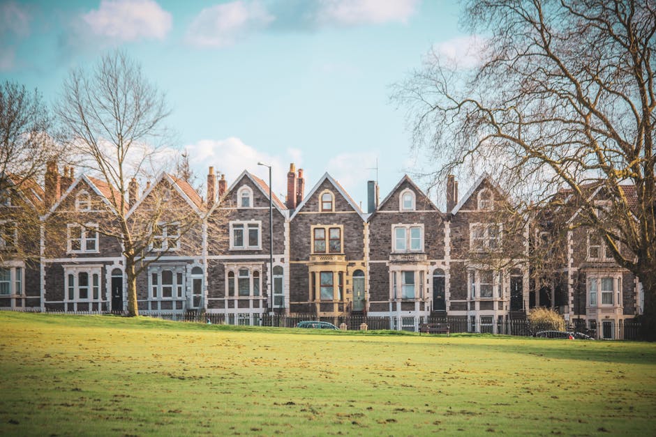 A row of historic stone-built terraced houses with pitched roofs, chimney stacks, and large sash windows, situated behind a well-maintained grassy park with a few leafless trees. The houses are set along a quiet residential street, with some parked cars visible in front. The image is taken during daylight under a partly cloudy sky, and the scene depicts a peaceful neighbourhood typical of urban house removals and relocation services near Bounds Green Station. Man with Van Bounds Green, a professional removals company, may assist with the logistics of home relocation, including furniture transport, packing, and loading processes, as evidenced by the spacious environment suitable for moving operations.