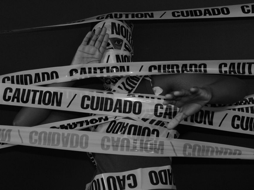 A person partially obscured behind multiple overlapping pieces of caution tape labeled 'CAUTION / CUIDADO' in bold black letters, with their left hand raised as if signaling stop and their right hand pointing forward. The individual is indoors, with a dark background and focused lighting highlighting their arms and the tape. The scene creates a visual impression of being restricted or warned, using safety tape as a central element. This imagery could relate to the careful handling and logistical planning involved in home relocations, furniture transport, or packing and moving services provided by companies like Man with Van Bounds Green, which specialises in removals in the Bounds Green area.