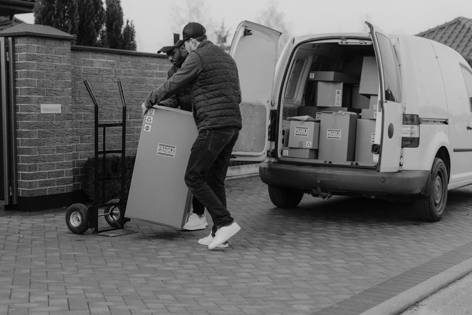A man with a beard and light skin, dressed in a blue and red windbreaker, black pants, and white sneakers with black and brown accents, is sitting on the rear bumper of a moving van with its back door