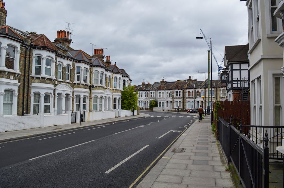 A quiet residential street on Bounds Green Road in BOUNDS GREEN lined with Victorian-style terraced houses, featuring bay windows, brick facades, and front gardens enclosed by black metal railings. The street has a paved pavement on the right side, with some patches of greenery and small trees. Parked cars are visible along the curb, and a person is walking on the sidewalk. Overhead, the sky is overcast with grey clouds, and there are construction cranes in the distance, indicating ongoing building work. The scene depicts a typical suburban environment suitable for home relocation or furniture transport activities, with no ongoing removals or moving equipment visible in this particular image, but it provides context for a neighbourhood where professional removals, such as those by Man with Van Bounds Green, might operate.