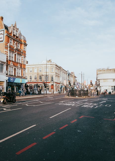 A wide view of a busy street in Bounds Green, Haringey, showing historic and modern multi-storey buildings lining the road. The scene includes parked cars and motorcycles along the curb, with a few pe