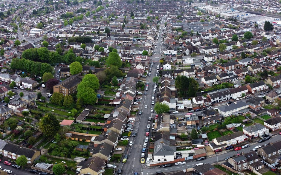 Aerial view of Bounds Green showing a row of mixed residential and commercial buildings lining a busy street, with cars and vans parked along the roadside. The scene includes a small roundabout with v