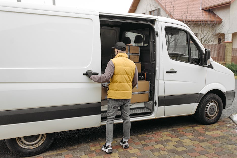 A man wearing a black cap, yellow and gray padded jacket, gray jeans, and sneakers is opening the sliding side door of a white delivery van parked on a residential street with brick paving. Inside the