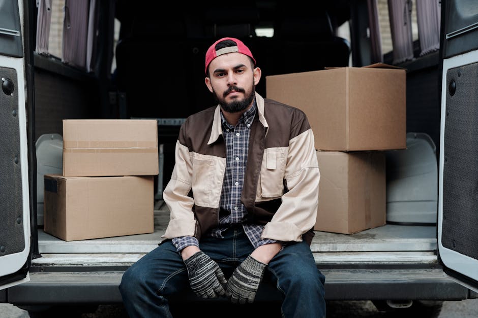A man wearing a black cap, yellow and gray padded jacket, gray jeans, and sneakers is opening the sliding side door of a white delivery van parked on a residential street with brick paving. Inside the