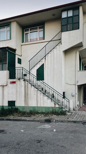 The exterior of a multi-storey residential building with a concrete facade painted in light beige, featuring green window frames and shutters. An external metal staircase with black railings runs along the side of the building, connecting the ground level to the first-floor entrance, which has a green door. The staircase is positioned close to a pavement made of concrete paving stones, with some scattered debris visible on the ground. Inside the building, through the open window on the upper floor, a glimpse of white curtains can be seen. The lower part of the building shows a basement window and a small external light fixture near the entrance. This setting illustrates typical challenges in home relocation and furniture transport when accessing apartments via narrow staircases, as managed by professional moving services like Man with Van Bounds Green, especially during detailed packing and loading processes involving multiple boxes and household items.