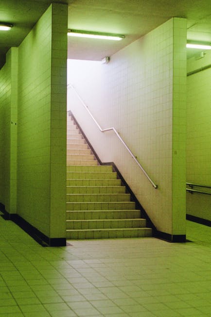 A set of tiled indoor stairs leading upwards in a narrow corridor with white tiled walls and a white handrail on the right side. The stairs are positioned centrally, with a slight turn at the top, and are illuminated by greenish fluorescent lighting from overhead fixtures. To the left of the stairs, a wall extends from the floor to the ceiling, partially obstructing the view of the upper landing. The floor adjacent to the stairs is also tiled with square light-colored tiles, and the overall environment appears to be part of a building’s communal or service area, such as a back entrance or stairwell. In the context of home relocation or furniture transport, this setting may be used for navigating between building floors during moving operations, which Man with Van Bounds Green handles professionally, especially when obstacles like narrow staircases pose challenges to the moving process.
