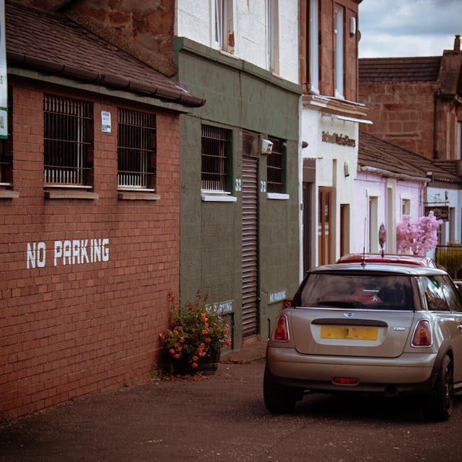 A narrow residential street scene showing parked vehicles and buildings with varied facades. In the foreground, a silver Mini Cooper with a UK license plate is parked close to a small sidewalk next to a brick wall with white 'No Parking' stenciling. Behind the car, a green-painted building with metal roller shutters and small barred windows is visible, suggesting a commercial or storage unit. To the right, a white building with a brown door and a sign partially visible indicates a business or office space. Further down the street, a pink-flowered tree adds color against a partly cloudy sky. Evidence of home relocation and moving logistics is implied by the parked vehicle and the urban environment, which may be used by Man with Van Bounds Green during property moves, furniture transport, and packing and loading activities. The scene captures a typical setting where parking restrictions are enforced in preparation for or during a house removal in the N11 area, with street parking suspensions and associated fines in effect.