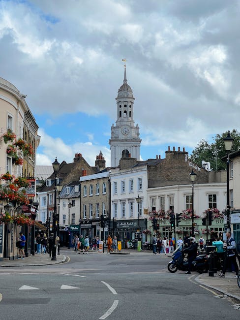 A busy street scene in Bounds Green featuring a row of multi-story buildings with retail shops on the ground level and residential or office spaces above. There are pedestrians walking on the sidewalks, some engaged in conversations or waiting, and others crossing the street. Several cars and a motorcycle are visible on the road, along with a few parked vehicles. The street is decorated with hanging flower baskets attached to lampposts on both sides. In the background, a tall, historic clock tower with a spire and weather vane rises prominently above the surrounding buildings under a partly cloudy sky, indicating a lively urban environment. This scene is captured during daylight hours, with natural lighting highlighting the architectural details, supporting context for house removals and furniture transport services by Man with Van Bounds Green.