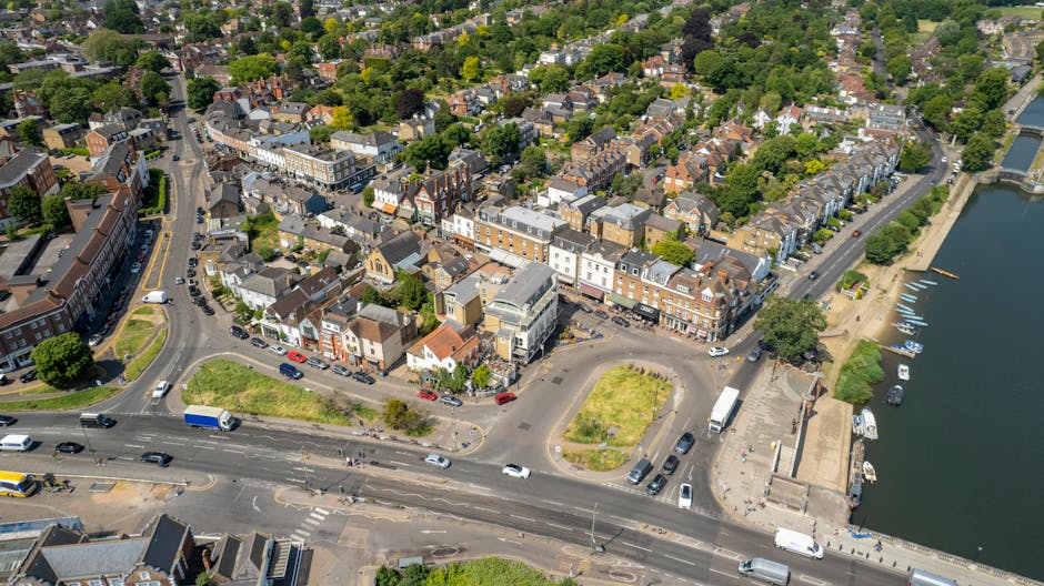 Aerial view of a residential area in Bounds Green showing a mix of terraced houses, apartment buildings, and retail shops along a main street. There are several parked cars lining the streets and a roundabout at the intersection. To the right, a river or lake with a paved promenade features boats docked along the shoreline. The sky is clear, and the area is lush with green trees and foliage. This image captures the urban environment where home relocations and furniture transport services by Man with Van Bounds Green may take place during packing, loading, and moving processes situated near the water and residential streets.