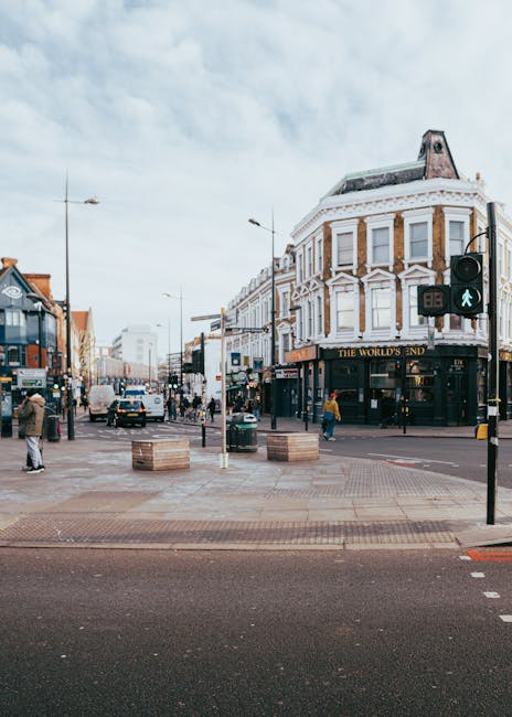 A busy street scene in Bounds Green featuring a row of multi-story buildings with retail shops on the ground level and residential or office spaces above. There are pedestrians walking on the sidewalks, some engaged in conversations or waiting, and others crossing the street. Several cars and a motorcycle are visible on the road, along with a few parked vehicles. The street is decorated with hanging flower baskets attached to lampposts on both sides. In the background, a tall, historic clock tower with a spire and weather vane rises prominently above the surrounding buildings under a partly cloudy sky, indicating a lively urban environment. This scene is captured during daylight hours, with natural lighting highlighting the architectural details, supporting context for house removals and furniture transport services by Man with Van Bounds Green.